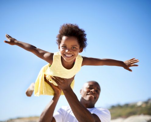 African American man lifting up a young African American girl as she smiles and stretches her arms as if she is an airplane